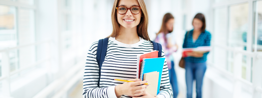 Marketing educacional: fotografia de uma estudante em um corredor de uma instituição de ensino. Ela está olhando para a câmera, sorrindo e segurando alguns livros.