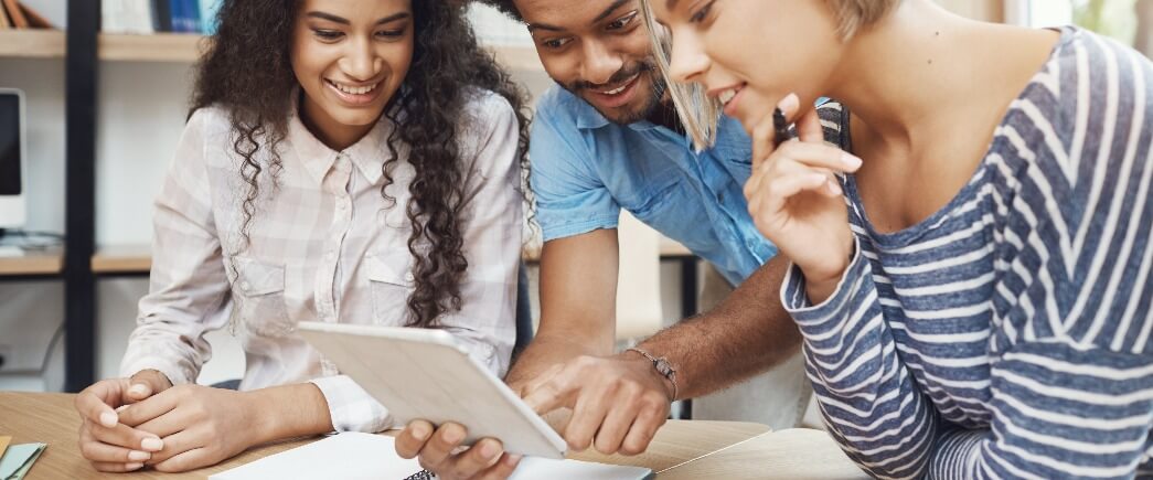 Como engajar alunos: fotografia de três alunos em uma biblioteca analisando algo em um tablet.