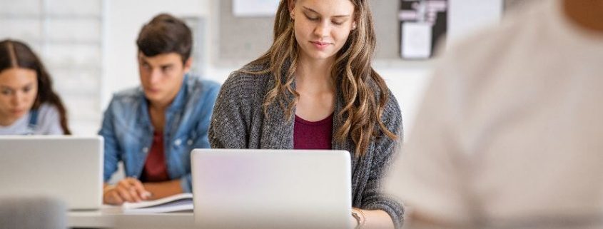 Universidade, faculdade ou centro universitário: fotografia de uma sala de aula com foco em uma mulher utilizando um computador.