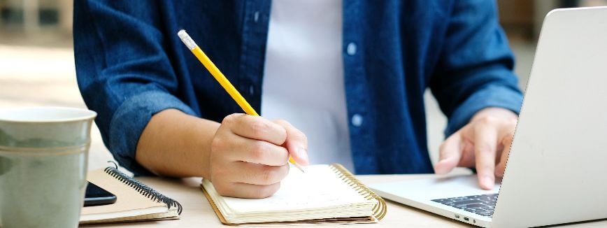Tipos de EaD: fotografia com foco em um estudante utilizando o computador para assistir aulas e fazendo anotações em um caderno.
