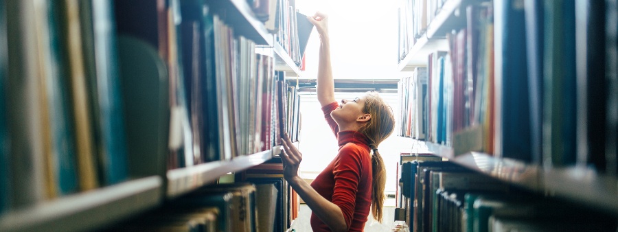 Catalogação de livros: fotografia de uma mulher organizando os livros nas estantes de uma biblioteca.