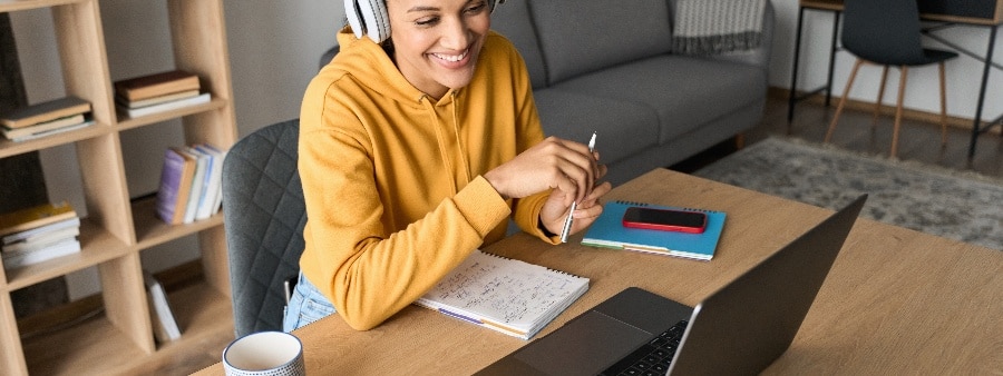 Ensino híbrido e metodologias ativas: fotografia de uma mulher sorrindo durante uma aula online. Ela está sentada à mesa, com um computador e materiais de estudo à sua frente.