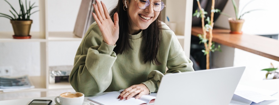 Pós-graduação lato sensu: fotografia de uma mulher assistindo a uma aula online. Ela sorr e está com uma das mãos levantada.