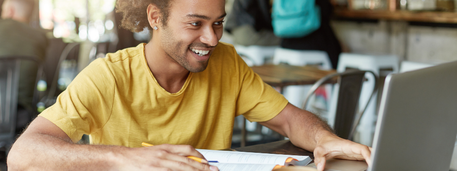 fotografia de um estudante sorrindo enquanto segura um lápis em uma mão e utiliza o computador.