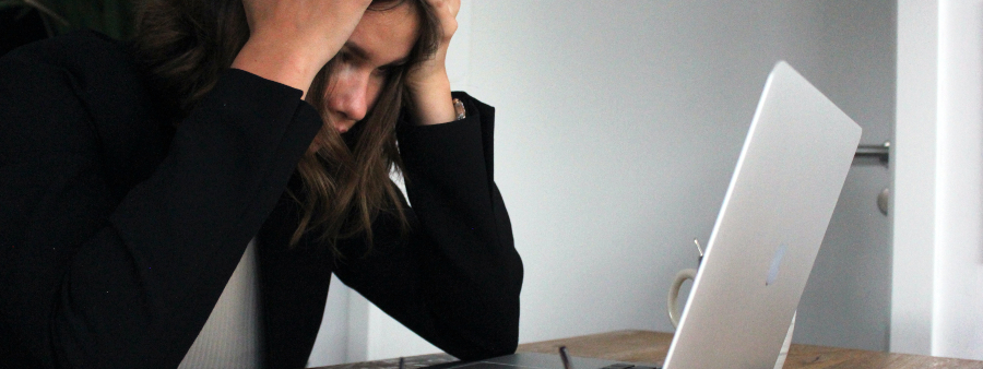 Síndrome de burnout em professores: fotografia de uma mulher com as mãos na cabeça em frente a um computador, expressando estar cansada e estressada.
